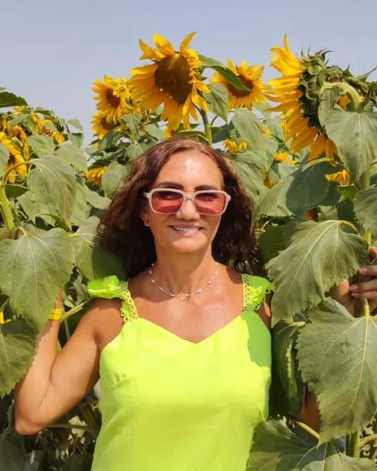 Francy Martins sorrindo em um campo de girassóis, com vestido verde-claro e óculos de lentes rosadas.